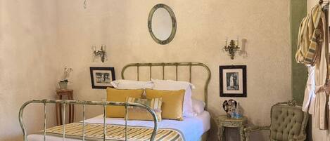 A bedroom with a metal bed frame, a chandelier, a round mirror, a small bedside table, a chair, and a patterned rug.