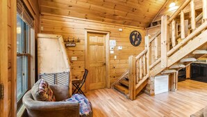 A wooden cabin interior with a staircase, a sofa with patterned cushions, a door, and a wall-mounted clock. - Southern Style - Hot Tub with TV, Game Room, Kids' Playhouse + Cornhole (Sevierville)