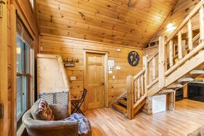 A wooden cabin interior with a staircase, a sofa with patterned cushions, a door, and a wall-mounted clock. - Southern Style - Hot Tub with TV, Game Room, Kids' Playhouse + Cornhole (Sevierville)
