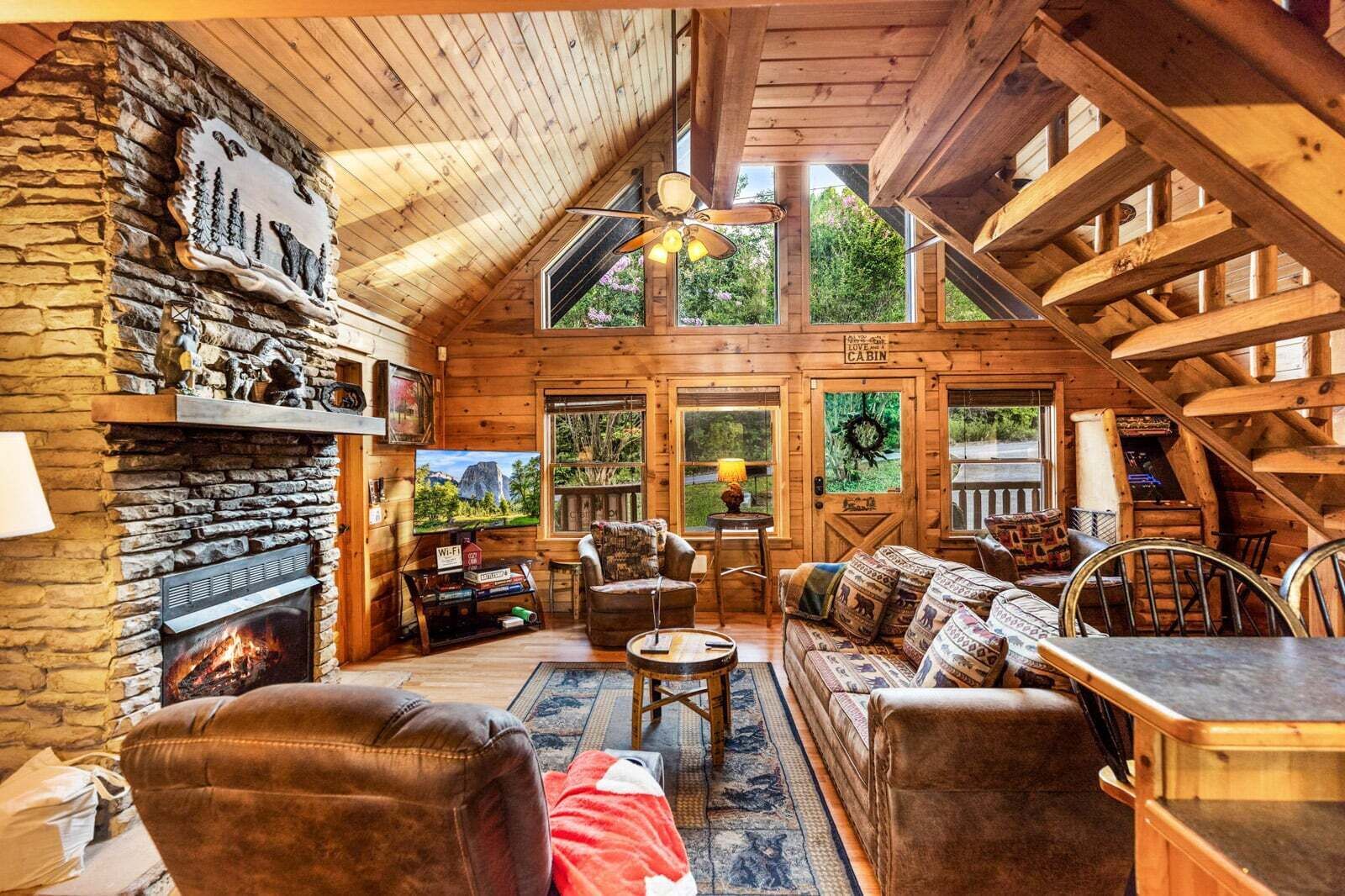 A log cabin interior with a stone fireplace, wooden furniture, and large windows.