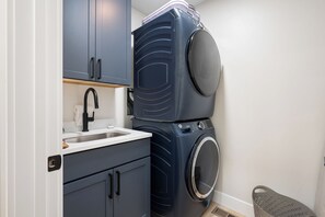 Een kleine wasruimte met een wasmachine en droger boven elkaar, een wasbak met kraan en kastjes.