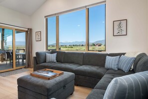 Ein modernes Wohnzimmer mit einem Ecksofa, einem Couchtisch und großen Fenstern mit Blick auf eine Wohngegend und Berge.