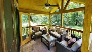 Eine überdachte Veranda mit Korbmöbeln, einem Deckenventilator und Blick auf ein bewaldetes Gebiet.