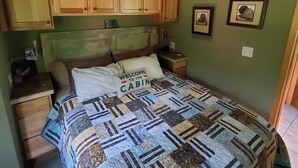 Ein Schlafzimmer im Blockhausstil mit Holzbalkendecke, einem Bett mit Quilttdecke, Holzschränken und Deckenventilator.