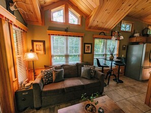 A cozy interior with a sofa, dining table, and kitchen area, featuring wooden beams and large windows.