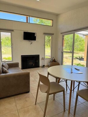 Una sala de estar con chimenea, un sofá, una mesa de comedor con sillas y ventanas amplias.