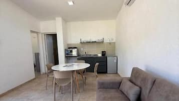 A living area with a sofa, a round table, and chairs. There is a kitchenette in the background.