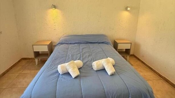A single bed with a blue bedspread and two white towels placed on it. There are two bedside tables, each with a lamp. The room has plain walls and a tiled floor.