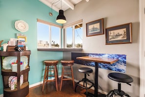 A room with a bay window, two stools, a table, and various framed pictures on the wall. - 1 Block to Beach | Charming 2BR Newport Bungalow w/ Patios, Bikes & Gear (Newport Beach)