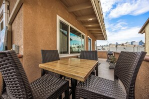 An outdoor patio with a wooden table and wicker chairs, a large window, and a view of the cityscape. - 1 Block to Beach | Charming 2BR Newport Bungalow w/ Patios, Bikes & Gear (Newport Beach)