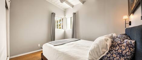 Um quarto com cama, cortinas, ventoinha no teto e mesa de cabeceira com abajur.