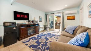 Ein Wohnzimmer mit Flachbildfernseher, der 'NETFLIX' anzeigt, einer Küchenzeile mit Kaffeemaschine und einem gemusterten Teppich.