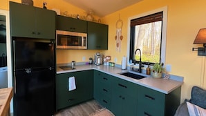 Een compacte keuken met een zwarte koelkast, een magnetron en een spoelbak. Er zijn kastjes in groentinten, en een raam met uitzicht op bomen.