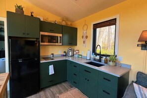 A compact kitchen with a black refrigerator, a microwave, and a sink. There are cabinets with a green color scheme, and a window with a view of trees. - Bison Farm - Vista Cabin (1 BR option) (Culpeper)