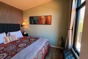 A hotel room with a bed, a wall-mounted painting, a bedside lamp, and a window with curtains. - Bison Farm - Vista Cabin (1 BR option) (Culpeper)