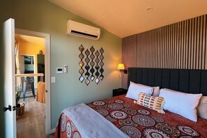 A hotel room with a bed, bedside table, wall-mounted air conditioner, decorative wall panel, and a door leading to a bathroom. - Bison Farm - Vista Cabin (1 BR option) (Culpeper)
