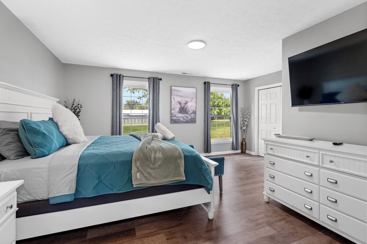 Quarto com cama, penteadeira, televisão e janela com cortinas.