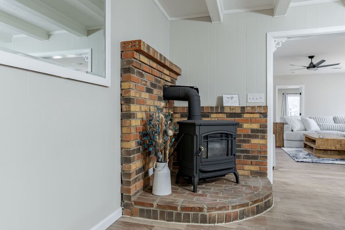 Una habitación con chimenea de ladrillo, estufa de leña negra y un jarrón blanco con flores secas.