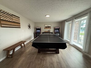 A room with a ping pong table, two arcade machines, a bench, and a large window with a view.