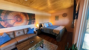 A cozy room with a wooden ceiling, a large abstract painting, a bed with a blue blanket, a brown leather sofa, and a small table with a remote control. - Bison Farm - Vista Cabin (2 BR option) (Culpeper)