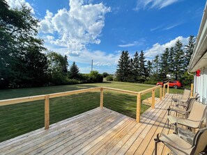 A wooden deck with a railing, outdoor furniture, and a view of a green lawn and trees. - BEAUTIFUL PRIVATE ACREAGE offers a 2-bedroom cottage in enchanting Cocagne (Cocagne)