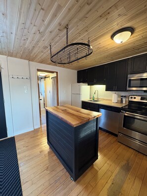 Una cocina con piso de madera, una isla central, gabinetes negros, electrodomésticos de acero inoxidable y una lámpara colgante.