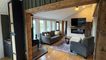 A spacious living area with a high ceiling, wooden beams, a fireplace, and a television mounted on a brick wall.