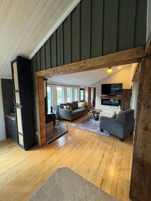 A spacious living area with a high ceiling, wooden beams, a fireplace, and a television mounted on a brick wall. - BEAUTIFUL PRIVATE ACREAGE offers a 2-bedroom cottage in enchanting Cocagne (Cocagne)