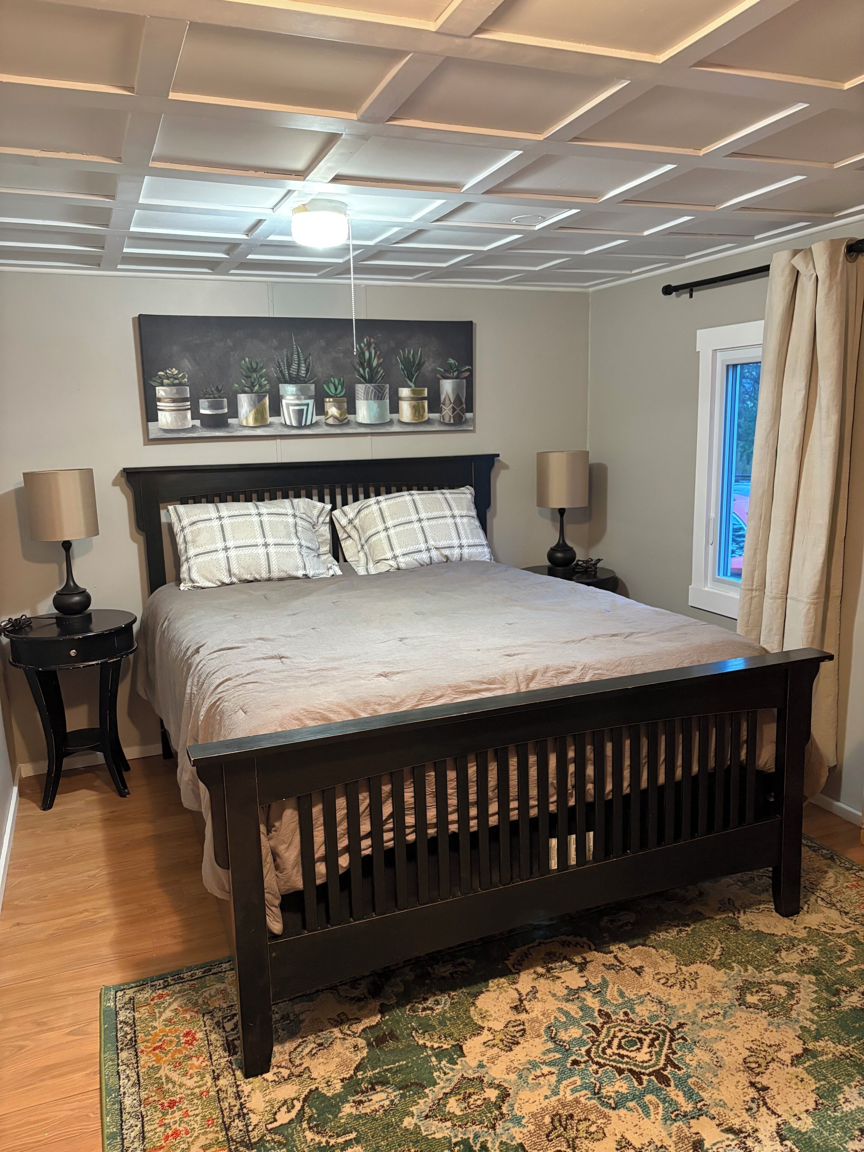 Une chambre à coucher comprenant un lit, deux tables de chevet, un luminaire au plafond, une fenêtre avec des rideaux et une étagère fixée au mur pour y poser des plantes.
