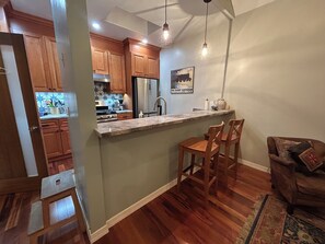 A kitchen with wooden cabinets, a stainless steel refrigerator, and a bar area with two wooden chairs. - Cozy cottage close to USC, downtown and quick trip to Culver City and Hollywood. (Los Angeles)