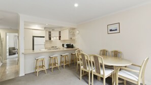A dining area with a round table and six chairs, a kitchen with a refrigerator and microwave, and a clock on the wall. - Osprey 1 - Queenscliff (QUEENSCLIFF)