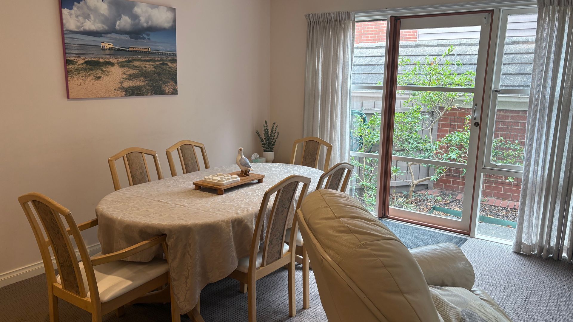 Comedor con una mesa dispuesta para seis personas, rodeada de sillas, un cuadro en la pared y vistas a través de un ventanal.