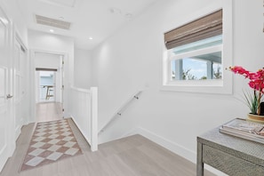 Un couloir avec un tapis à carreaux, une fenêtre avec des stores, un escalier et une porte donnant sur une autre pièce.