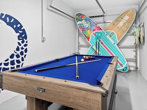 A pool table with a blue felt surface, pool cues, and balls, and a surfboard with a colorful design leaning against a wall.