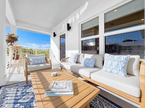 A sunlit outdoor seating area with a wooden table, a sofa with patterned cushions, and a view of the sky.