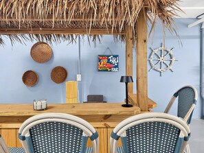 Une chambre décorée sur le thème de la plage, avec une table en bois, des chaises en osier, un toit de chaume et une enseigne indiquant « TIKI BAR ».
