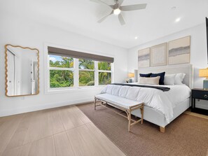A bedroom with a bed, bedside tables, a bench, a ceiling fan, and a window with a view of greenery.