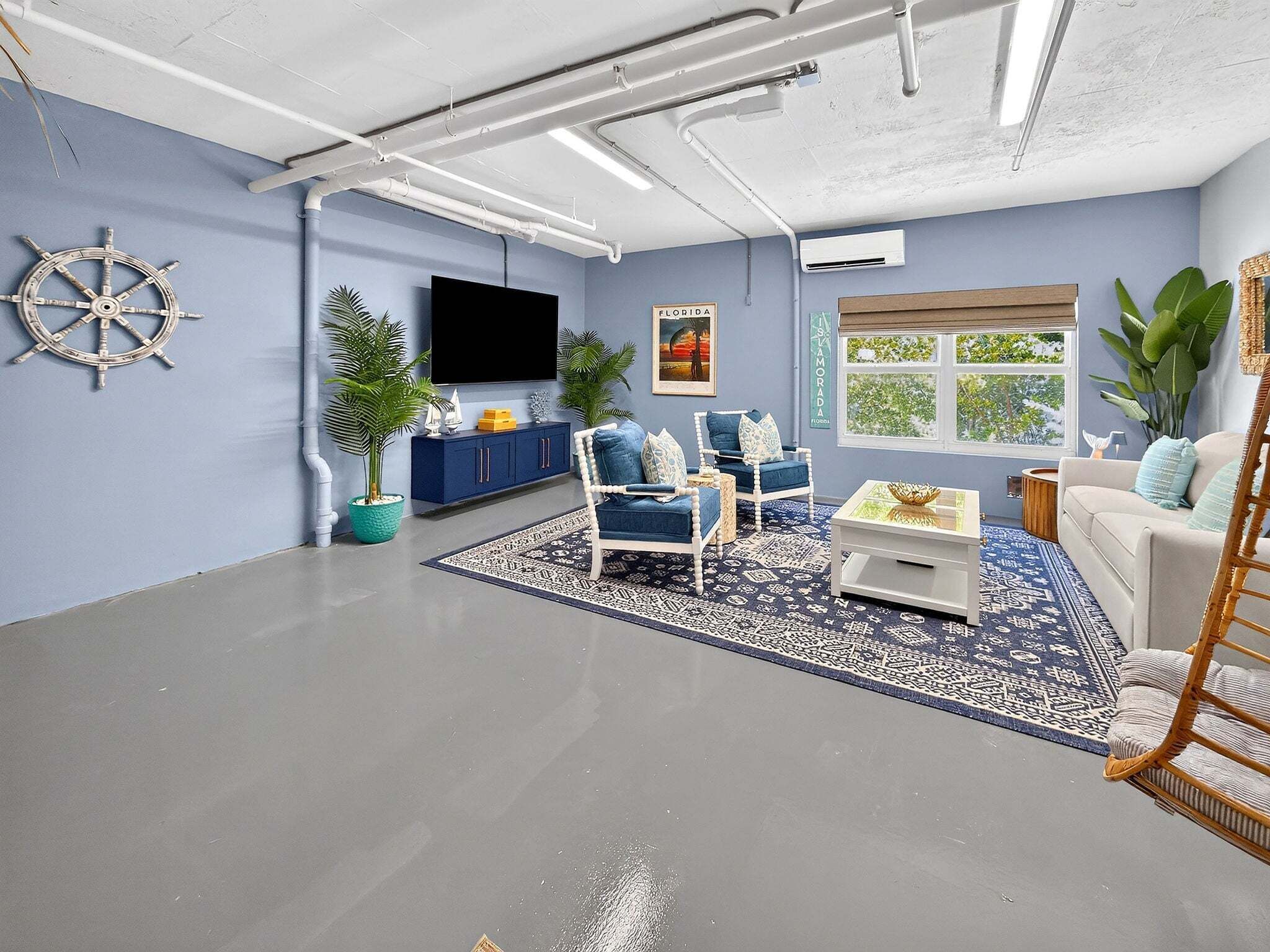 A living room with a flat-screen TV, a sail wheel decoration, a Florida poster, a blue sofa, a white armchair, and a large window with a view of trees.