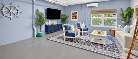 A living room with a flat-screen TV, a sail wheel decoration, a Florida poster, a blue sofa, a white armchair, and a large window with a view of trees.