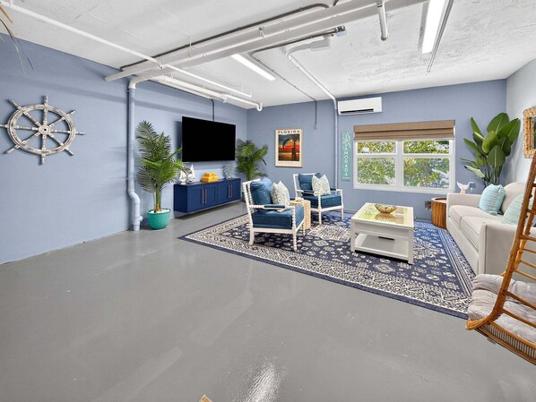 A living room with a flat-screen TV, a sail wheel decoration, a Florida poster, a blue sofa, a white armchair, and a large window with a view of trees.