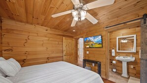 A wooden room with a bed, a ceiling fan, a TV, and a bathroom with a sink and mirror. - Spacious Mountain Getaway Minutes from Boone (Boone)