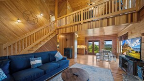 A spacious living area with a high ceiling, wooden beams, a blue sofa, a round coffee table, and a television displaying a scenic landscape. - Mountain Retreat with Long-Range Views Near Boone (Boone)