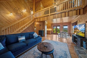 A spacious living area with a high ceiling, wooden beams, a blue sofa, a round coffee table, and a television displaying a scenic landscape. - Mountain Retreat with Long-Range Views Near Boone (Boone)