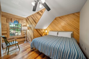 A bedroom with a sloped ceiling, wooden walls, a bed with striped bedding, a nightstand with a lamp, a chair, and a window with a view of greenery. - Mountain Retreat with Long-Range Views Near Boone (Boone)