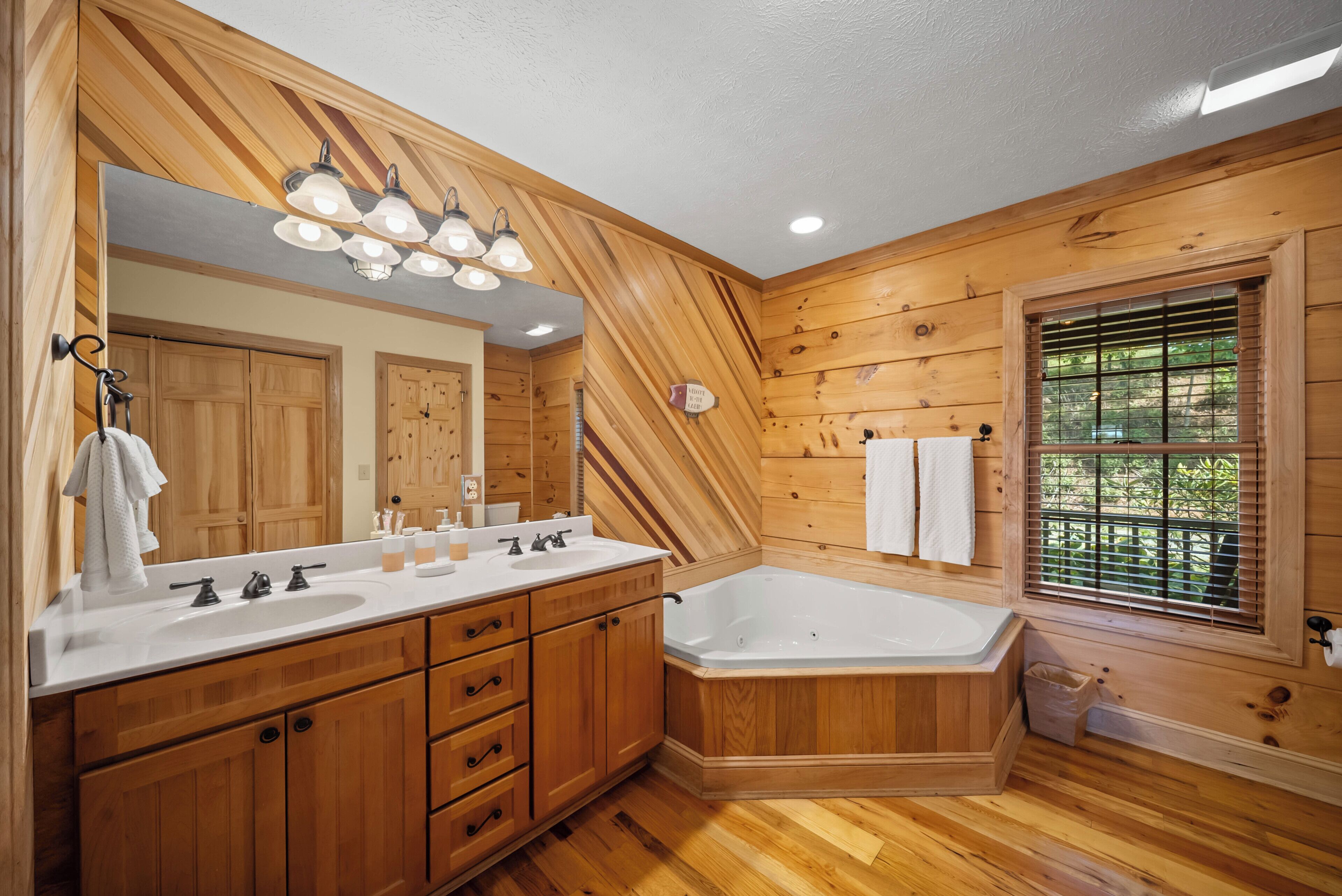Ein Badezimmer aus Holz mit einem großen Spiegel, einem Doppelwaschtisch, einer Badewanne und einem Fenster mit Jalousien.