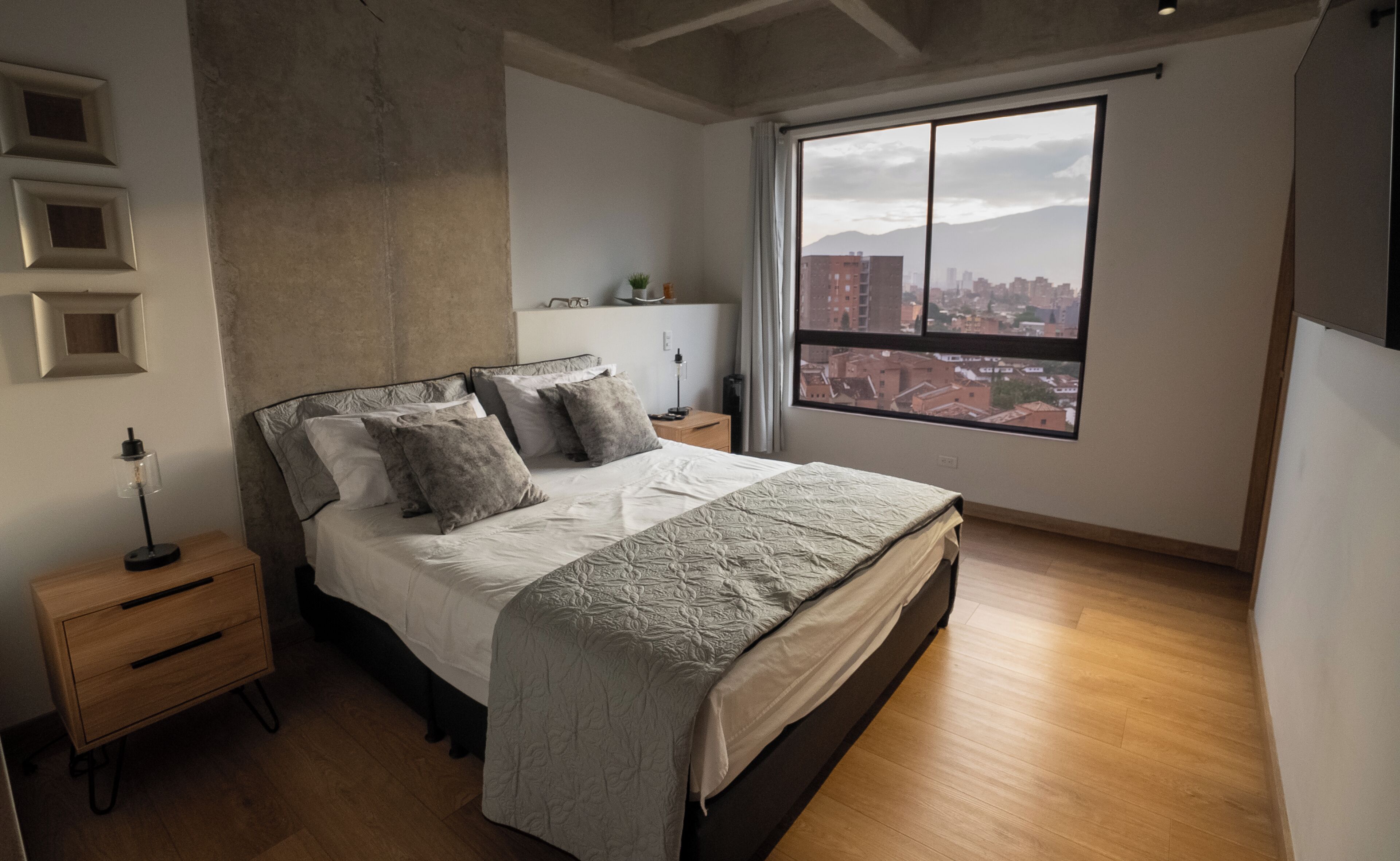 A bedroom with a large bed, a nightstand, a lamp, and a window with a city view.