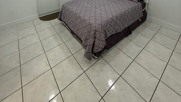 Ein Schlafzimmer mit Bett, einer Tür, einem Fenster mit Jalousien, einer Klimaanlage und Fliesenboden.