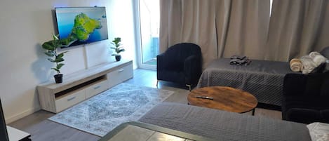 A modern living room with a flat-screen TV mounted on the wall, a sofa, a coffee table, and a bed with a quilted cover.