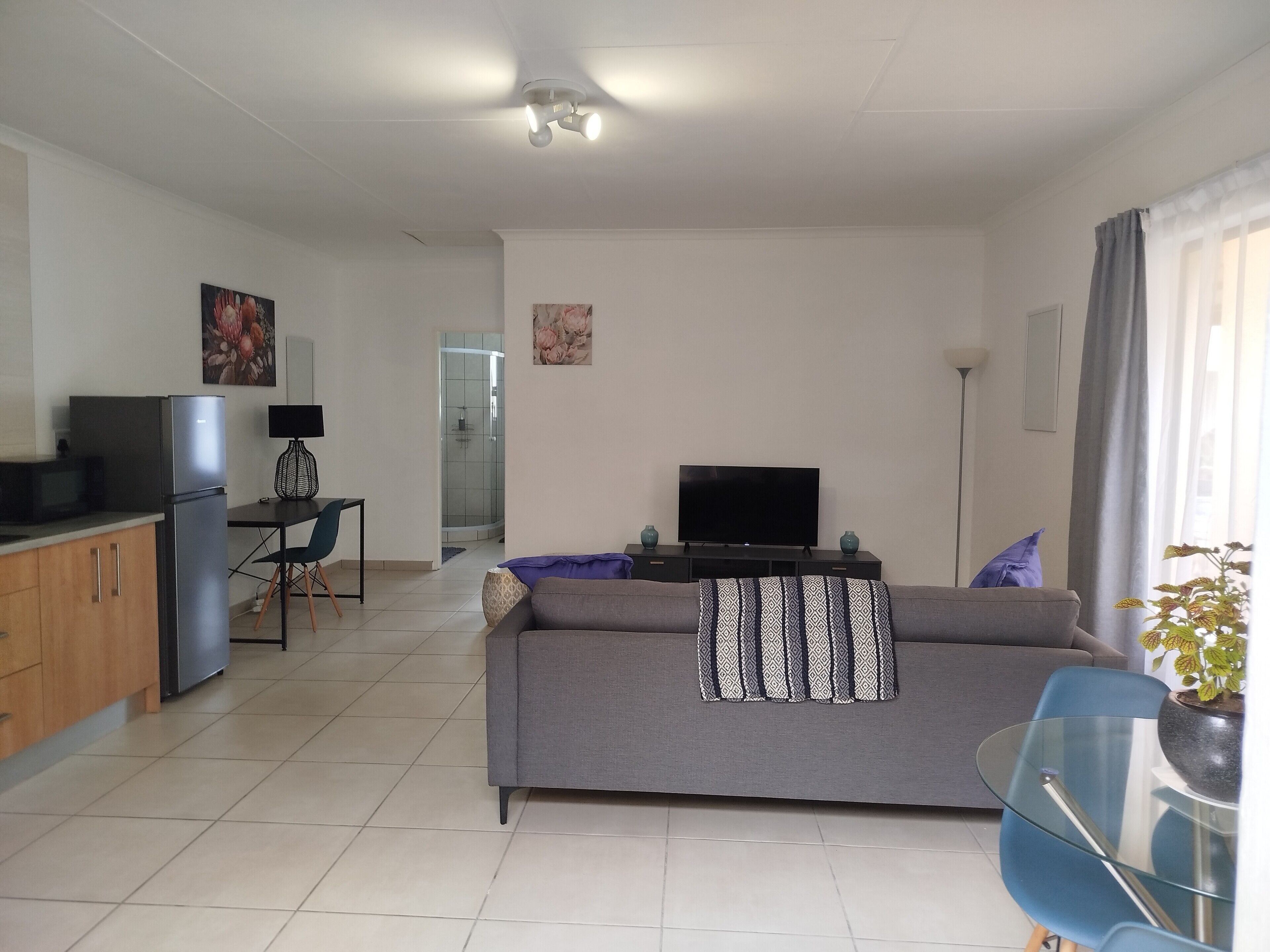 A modern living room with a sofa, a flat-screen TV, a dining area with a table and chairs, and a kitchen with a refrigerator.