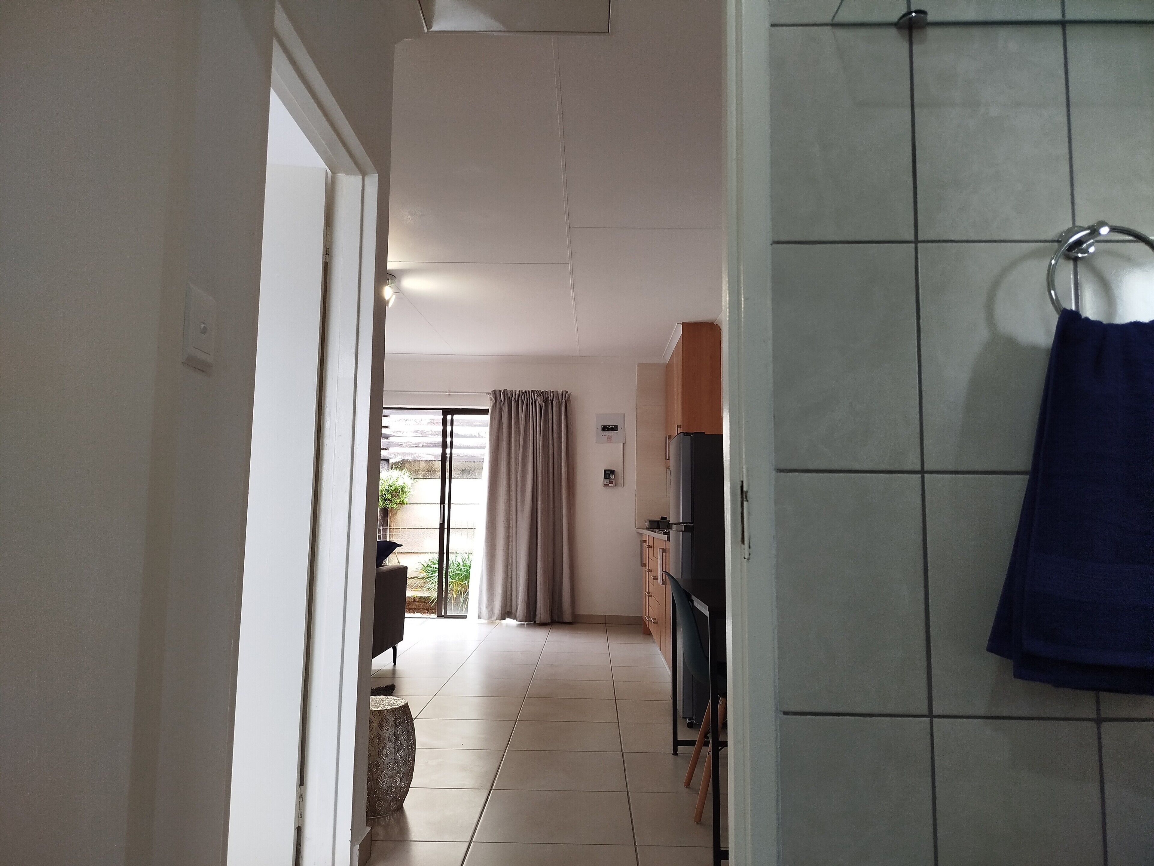 Un couloir mène à une cuisine équipée d’un réfrigérateur, d’un évier et d’une fenêtre avec des rideaux.